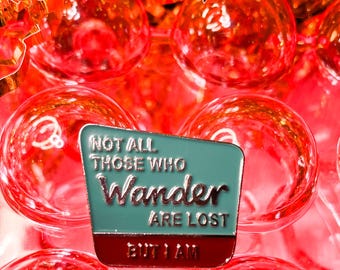 May include: A teal blue enamel pin with a silver border and white text that reads "Not all those who wander are lost but I am". The pin is on a red background.