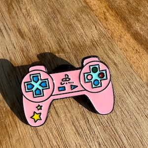 Puede incluir: Un pin de esmalte rosa con forma de mando de videojuego con botones azules y verdes. El mando tiene dos estrellas amarillas en el lado izquierdo.