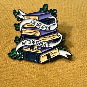 Peut inclure: Une épingle en émail colorée représentant une pile de livres avec une bannière qui dit "Ask Me About My To Be Read Pile". Les livres sont violets, beiges et vert foncé, avec des feuilles vertes et une bannière blanche. L'épingle est posée sur un fond jaune.
