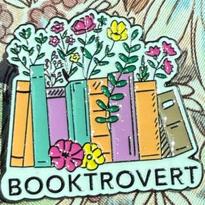 Peut inclure: Épinglette colorée en émail représentant une pile de livres avec des fleurs qui en sortent. L'épingle a une bordure vert menthe et le mot "BOOKTROVERT". Les livres sont orange, verts et violets.
