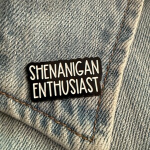 May include: A rectangular enamel pin with a black background and a scalloped edge. The pin features the white text "SHENANIGAN ENTHUSIAST" in a bold, sans-serif font. The pin is attached to a light blue denim fabric with visible stitching.
