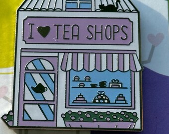 May include: Enamel pin depicting a tea shop with the text "I ♥ TEA SHOPS". The shop features a purple awning, a window display of tea items, and a black cat on the roof. The pin is white, purple, and blue.