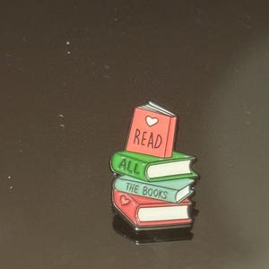Peut inclure: Une épingle en émail colorée représentant une pile de livres. Le livre du dessus est rouge avec le mot "READ" et un cœur. Le livre du milieu est vert avec "ALL" et le livre du bas est bleu avec "THE BOOKS".