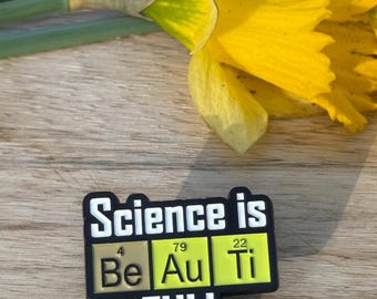 May include: A black enamel pin with the text "Science is Be Au Ti FULL" in white lettering. The letters "Be", "Au", and "Ti" are in yellow squares with the atomic numbers 4, 79, and 22 above them.