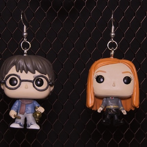May include: A pair of novelty earrings featuring miniature figurines of characters from a popular fantasy series. One earring depicts a boy with glasses, the other a girl with red hair. The earrings hang from silver-colored hooks.