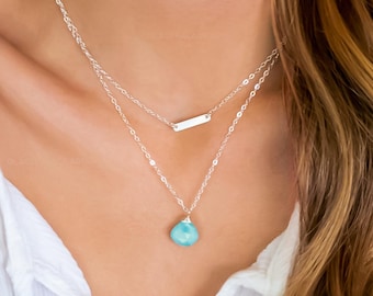May include: A silver necklace with a turquoise pendant and a small silver bar charm. The necklace is layered and has a delicate chain.