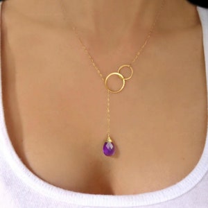 May include: A gold-colored necklace with two interlocking circles and a dangling purple gemstone pendant. The necklace is worn against a white top. The text "Glass Palace Arts" is visible at the top.
