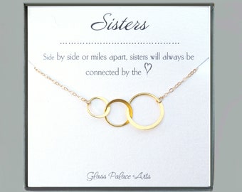 May include: Gold necklace with three interlocking circles and a card that reads "Sisters, Side by side or miles apart, sisters will always be connected by the heart. Glass Palace Arts."