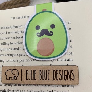 May include: A green avocado bookmark with a brown pit, a black mustache, and two black eyes. The bookmark has the text "ELLIE BLUE DESIGNS" on a brown rectangular tag.