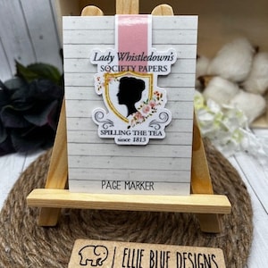 May include: A white page marker with a pink magnetic strip. The design features a silhouette of a woman in a floral frame with the text "Lady Whistledown's Society Papers, Spilling the Tea since 1813, Page Marker".