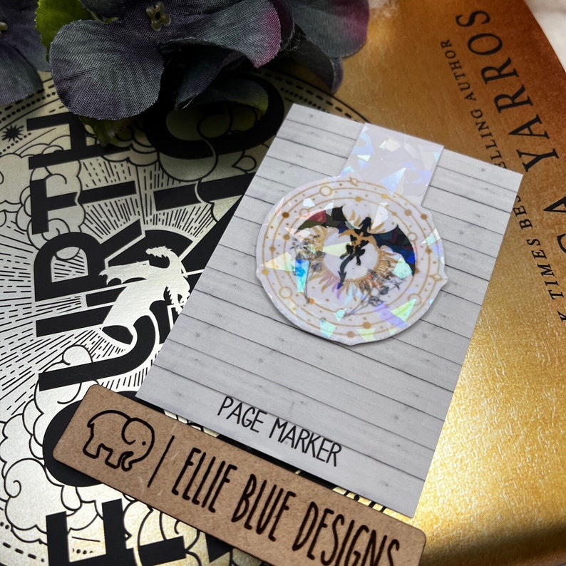 May include: A white page marker with a holographic sticker of a black dragon in a circle with gold accents. The text "PAGE MARKER" and "ELLIE BLUE DESIGNS" is printed on the page marker.