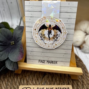May include: A white and gold circular sticker with a black dragon silhouette in the center. The sticker has a holographic finish and says "Page Marker" at the bottom.