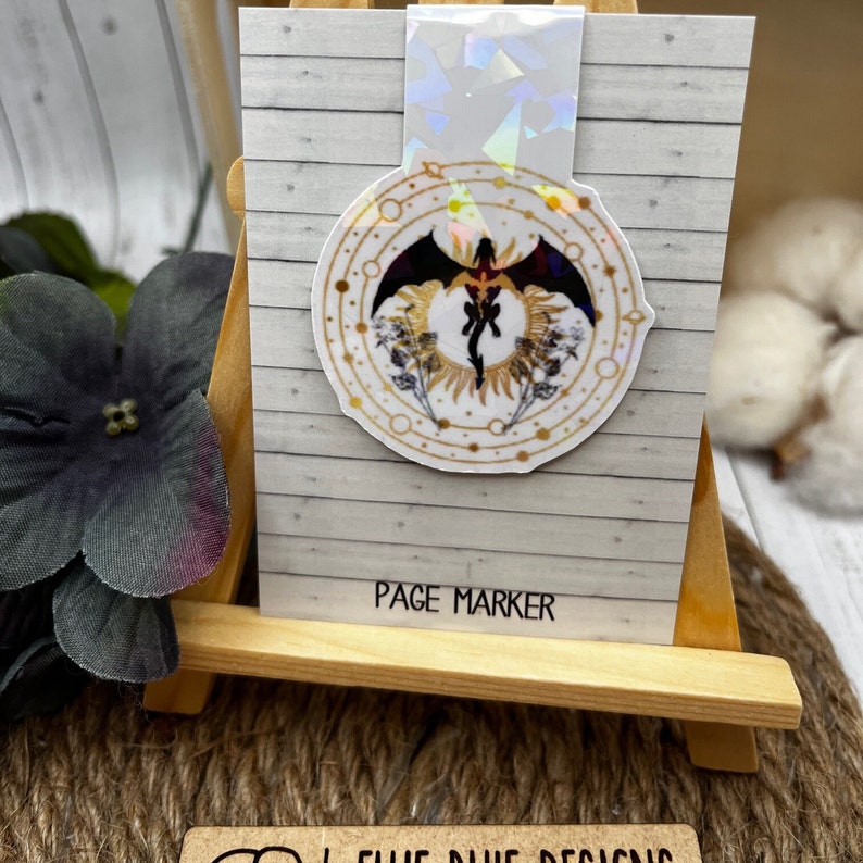 May include: A white and gold circular sticker with a black dragon silhouette in the center. The sticker has a holographic finish and says "Page Marker" at the bottom.
