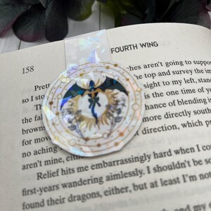 May include: A gold and white circular bookmark with a black dragon design and the words "FOURTH WING" printed on the page behind it.