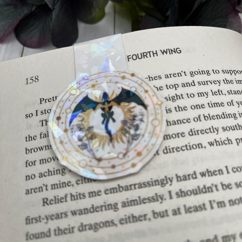 May include: A gold and white circular bookmark with a black dragon design and the words "FOURTH WING" printed on the page behind it.