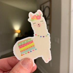 May include: A white llama sticker with a colorful patterned skirt and a flower crown. The llama has closed eyes and a pink nose.