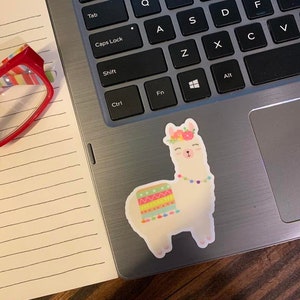 May include: A white llama sticker with a colorful floral headband, a rainbow patterned blanket, and a string of colorful beads around its neck. The sticker is on a silver laptop.