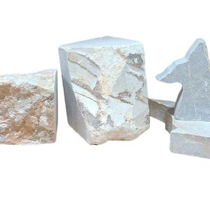 Può includere: Una collezione di sculture in pietra di forma irregolare, nei toni del grigio, beige e marrone. I pezzi variano per dimensioni e forma, inclusi blocchi rettangolari e forme astratte. La texture naturale della pietra è visibile.