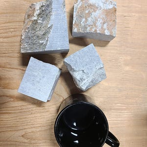 Pu&ograve; includere: Quattro blocchi di pietra irregolari e grezzi con superfici grigie e marroni, disposti vicino a una tazza in ceramica nera. Le pietre presentano un mix di facce lisce e piatte e bordi frastagliati e naturali, con texture variegate.
