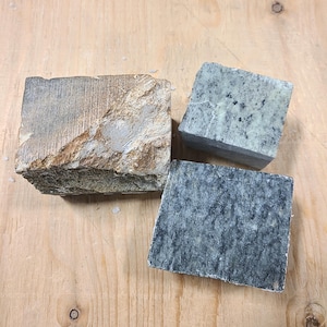 Pu&ograve; includere: Tre blocchi di pietra rettangolari con texture e colori vari. Un blocco &egrave; marrone e beige con una superficie ruvida e irregolare. Un secondo blocco &egrave; grigio chiaro con macchie scure. Il terzo blocco &egrave; grigio scuro con venature.