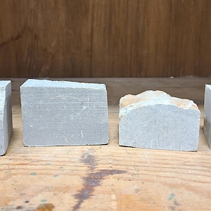 Pu&ograve; includere: Quattro blocchi di pietra grigia con diverse trame e forme sono disposti su una superficie di legno. I blocchi hanno bordi ruvidi e naturali e una tavolozza di colori tenui, che suggerisce un'estetica grezza e organica.