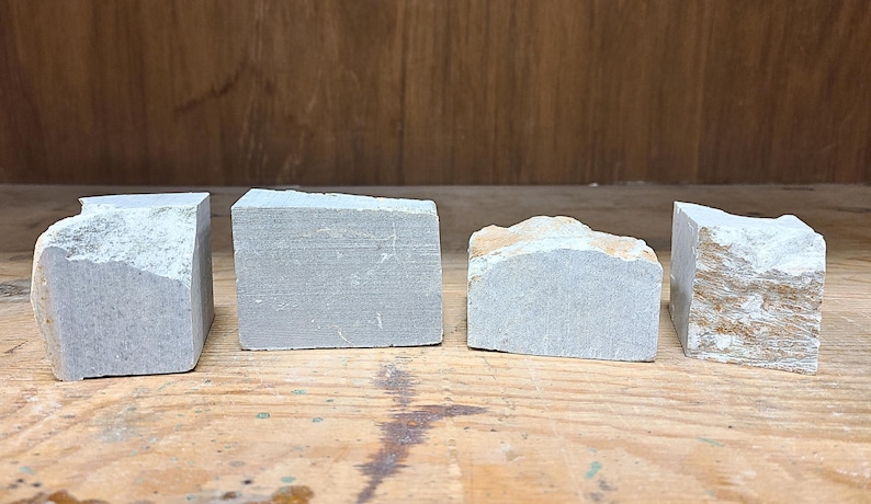 Pu&ograve; includere: Quattro blocchi di pietra grigia con diverse trame e forme sono disposti su una superficie di legno. I blocchi hanno bordi ruvidi e naturali e una tavolozza di colori tenui, che suggerisce un'estetica grezza e organica.