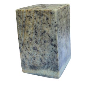 Può includere: Una saponetta rettangolare con una superficie macchiettata e strutturata. Il sapone ha una palette di colori beige, grigio e nero, con una sottile sfumatura blu su un lato. Il sapone è un blocco solido, con un fondo leggermente arrotondato.