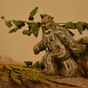 May include: A carved, olive-green figurine of Bigfoot with white etched details. The sculpture is posed in a walking stance and is set on a piece of wood with greenery. The background is a neutral tan color.