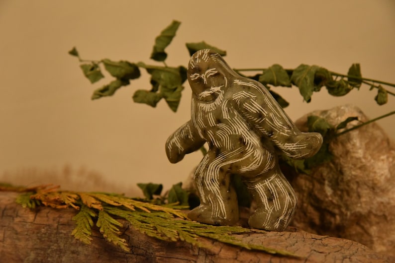 May include: A carved, olive-green figurine of Bigfoot with white etched details. The sculpture is posed in a walking stance and is set on a piece of wood with greenery. The background is a neutral tan color.