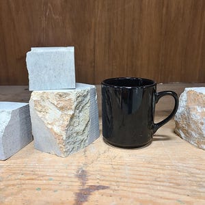 Pu&ograve; includere: Una tazza in ceramica nera con manico &egrave; posizionata vicino a diversi blocchi di pietra grigio chiaro di forma irregolare con accenti beige e marroni. La tazza ha una finitura lucida. Lo sfondo &egrave; una superficie di legno.