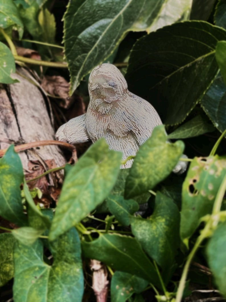 May include: A small, light-colored statue of a creature, possibly a sloth or similar animal, is nestled among green leaves. The statue has a textured surface and is positioned on a piece of wood. The leaves are a mix of dark and light green.