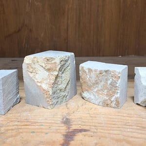 Pu&ograve; includere: Quattro blocchi di pietra dalla forma irregolare, nei toni del grigio e del beige, disposti su una superficie di legno. I blocchi hanno forme irregolari e superfici ruvide, suggerendo pietra naturale.