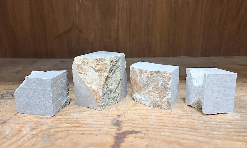 Pu&ograve; includere: Quattro blocchi di pietra dalla forma irregolare, nei toni del grigio e del beige, disposti su una superficie di legno. I blocchi hanno forme irregolari e superfici ruvide, suggerendo pietra naturale.