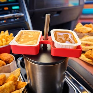 Puede incluir: Un soporte rojo para salsas con dos compartimentos, que contiene pequeños recipientes de salsas para mojar, se encuentra sobre un vaso de acero inoxidable. El soporte tiene una pajita plateada en el centro. Se ven patatas fritas y nuggets de pollo en el fondo.
