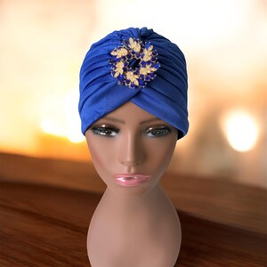 May include: A royal blue turban with a decorative jeweled brooch. The brooch features a circular design with gold-toned leaves and blue and white gemstones. The turban is displayed on a mannequin head.