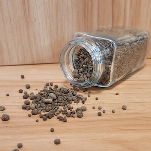May include: A glass jar with a clear lid is tipped over, spilling out small, dark brown, round pellets onto a light brown wooden surface.