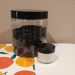 May include: Two clear plastic jars with black lids. One large jar is filled with dark, irregularly shaped objects. A smaller jar contains a white powder. The jars are on a white and orange patterned surface.