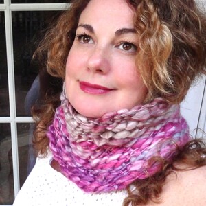 May include: A woman with curly brown hair is wearing a pink and white knitted infinity scarf. The scarf is made with a chunky yarn and has a soft, textured look.