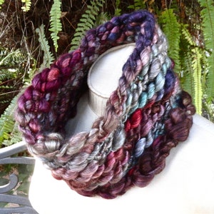 Op de afbeelding: Een dikke gebreide cowl-hals sjaal in tinten van paars, grijs, teal en bruin. De sjaal is gemaakt met een dik garen en heeft een textuur.