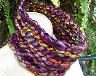 May include: A thick, colorful knitted neck warmer in shades of purple, gold, and brown. The neck warmer is made with a chunky yarn and has a textured, woven look.