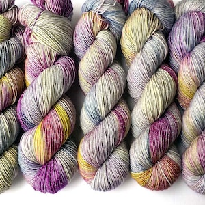 May include: Five skeins of yarn in a variegated colorway with shades of purple, blue, yellow, and grey. The yarn is a fingering weight and has a subtle sheen.
