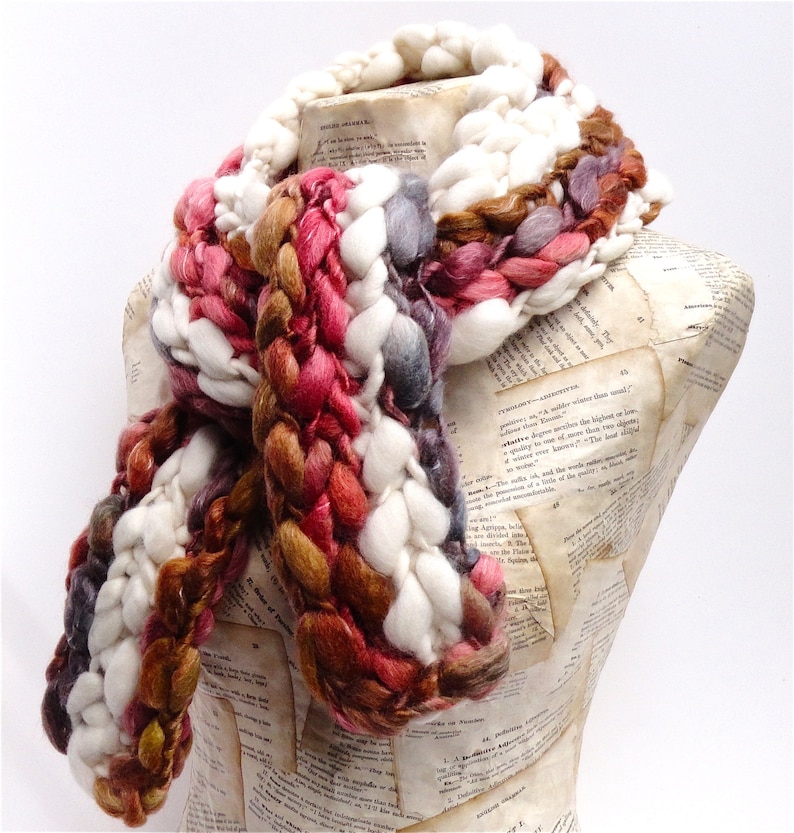 May include: A chunky knit scarf in white, brown, pink, and grey. The scarf is made of soft yarn and has a unique braided design.