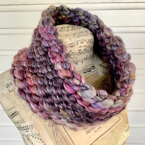 May include: A thick, chunky knit cowl in shades of purple, pink, and gray. The cowl is made with a soft, textured yarn and is perfect for keeping warm in the winter.
