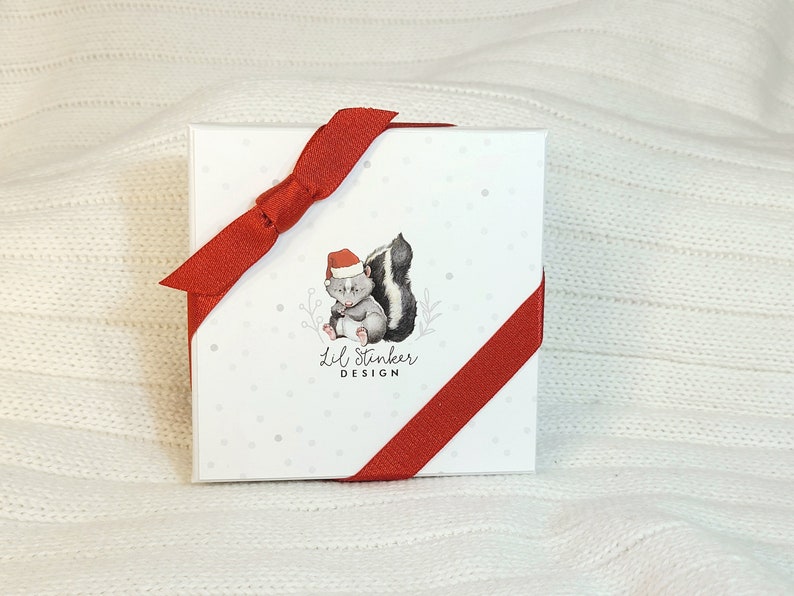 Pu&ograve; includere: Una scatola regalo bianca con un nastro e un fiocco rossi. La scatola ha uno sfondo bianco con pois grigi e un'illustrazione di un fumetto di una puzzola che indossa un cappello di Babbo Natale. Il testo "Lil Stinker Design" &egrave; stampato sotto l'illustrazione.