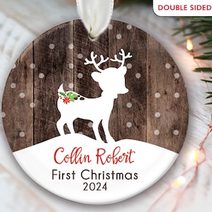 K&ouml;nnte beinhalten: Ein runder, h&ouml;lzerner Weihnachtsbaumschmuck mit einer wei&szlig;en Silhouette eines Rentiers mit einer roten Schleife und gr&uuml;nem Stechpalmenzweig. Der Schmuck hat einen wei&szlig;en Hintergrund mit dem Text "Collin Robert First Christmas 2024" in Rot.
