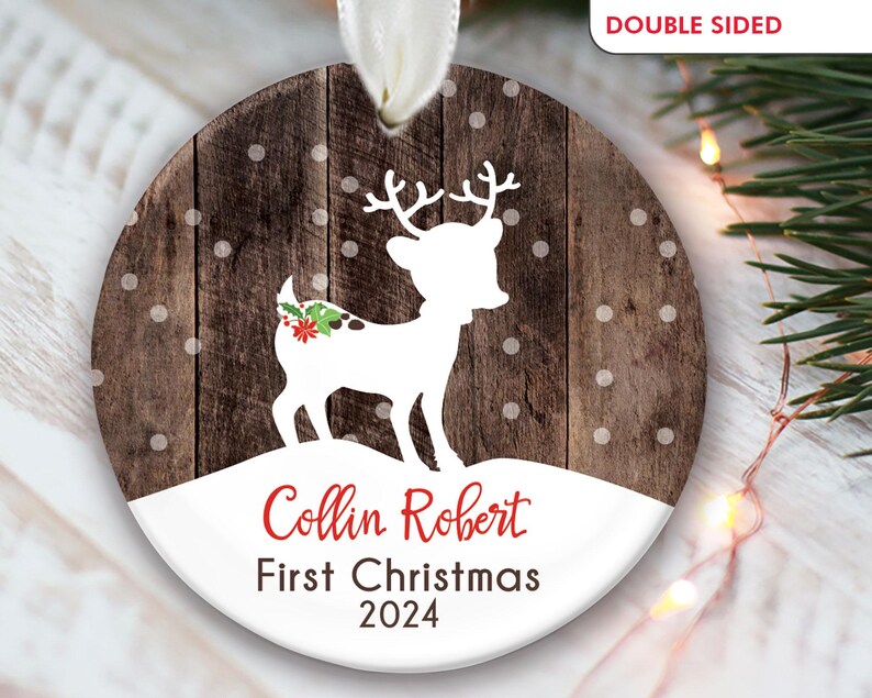 K&ouml;nnte beinhalten: Ein runder, h&ouml;lzerner Weihnachtsbaumschmuck mit einer wei&szlig;en Silhouette eines Rentiers mit einer roten Schleife und gr&uuml;nem Stechpalmenzweig. Der Schmuck hat einen wei&szlig;en Hintergrund mit dem Text "Collin Robert First Christmas 2024" in Rot.