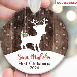 K&ouml;nnte beinhalten: Ein runder, wei&szlig;er, h&ouml;lzerner Weihnachtsbaumschmuck mit einer wei&szlig;en Silhouette eines Rentiers mit einer roten Schleife. Der Schmuck hat einen braunen Holzmaserungshintergrund mit wei&szlig;en Punkten. Der Text "Sean Malcolm First Christmas 2024" ist in Rot auf den Schmuck gedruckt.
