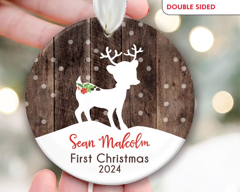 K&ouml;nnte beinhalten: Ein runder, wei&szlig;er, h&ouml;lzerner Weihnachtsbaumschmuck mit einer wei&szlig;en Silhouette eines Rentiers mit einer roten Schleife. Der Schmuck hat einen braunen Holzmaserungshintergrund mit wei&szlig;en Punkten. Der Text "Sean Malcolm First Christmas 2024" ist in Rot auf den Schmuck gedruckt.