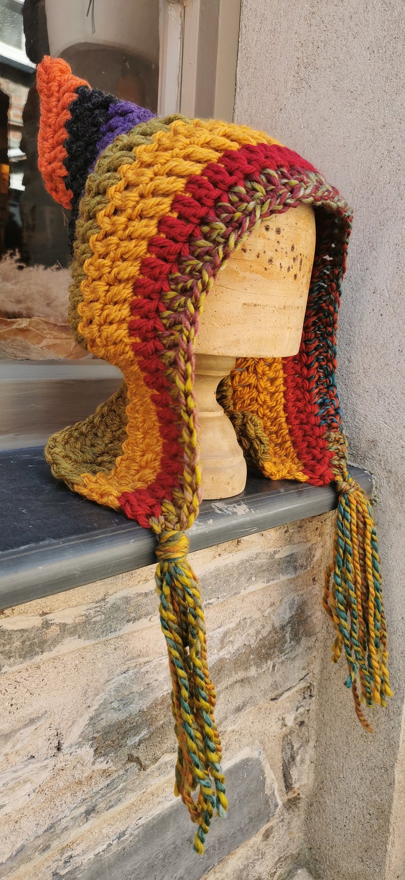 K&ouml;nnte beinhalten: Eine bunte H&auml;kelm&uuml;tze mit einer langen, spitzen Spitze und Quasten. Die M&uuml;tze ist in verschiedenen Farben gefertigt, darunter Rot, Orange, Gelb, Gr&uuml;n und Lila.