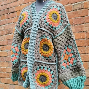 May include: A crocheted cardigan in a blue, brown and orange colour scheme. The cardigan is made of granny squares with a floral pattern. The cardigan is worn on a mannequin with a crocheted hat.